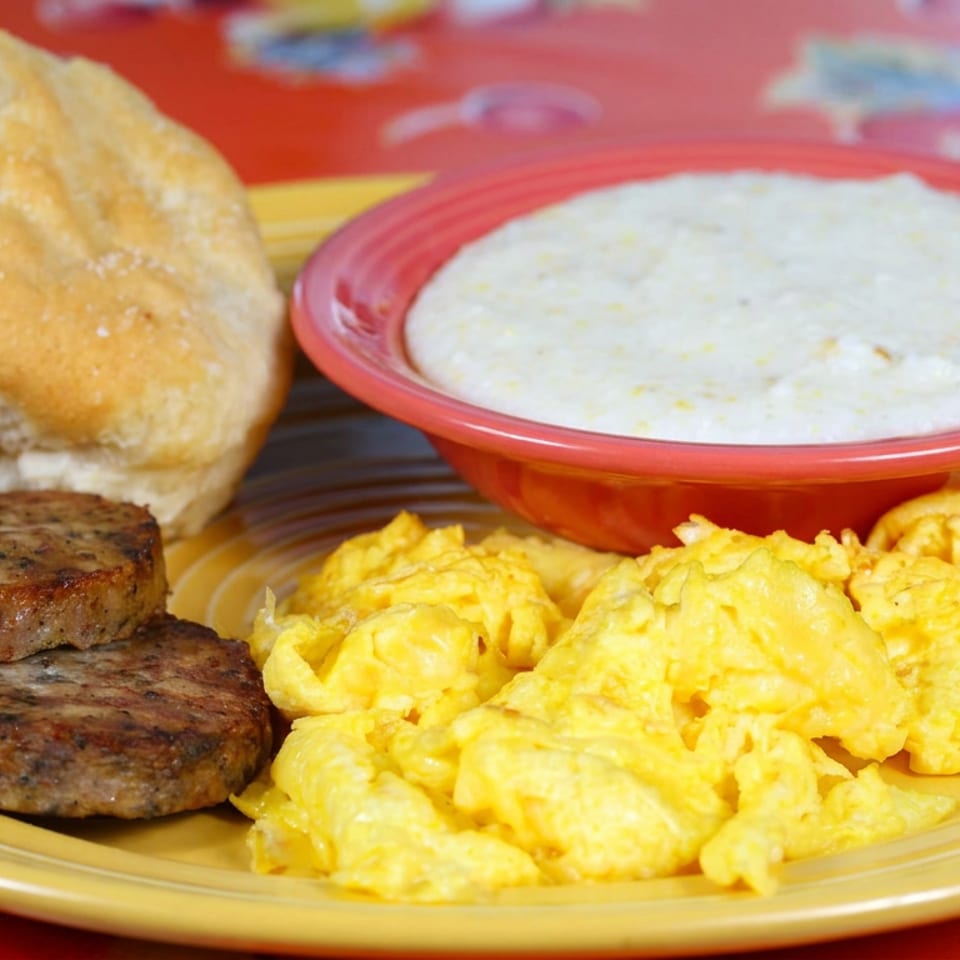 Breakfast plate with scrambled eggs, two sausage patties, toasted bread, and creamy sauce in a red bowl.