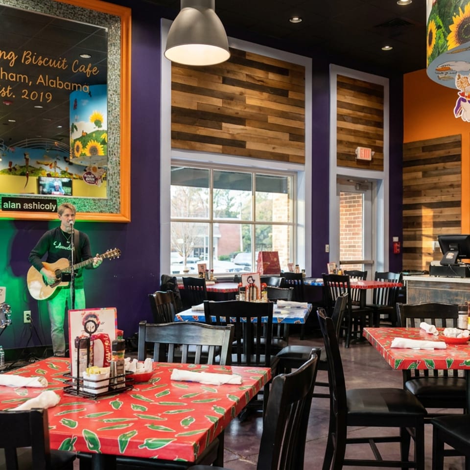 Colorful Flying Biscuit Cafe dining room with red tables, black chairs, open kitchen, and musician performing guitar.