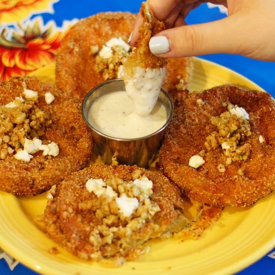 Four golden fried patties topped with crumbled cheese surround creamy dipping sauce on a yellow plate, while a hand dips one patty.