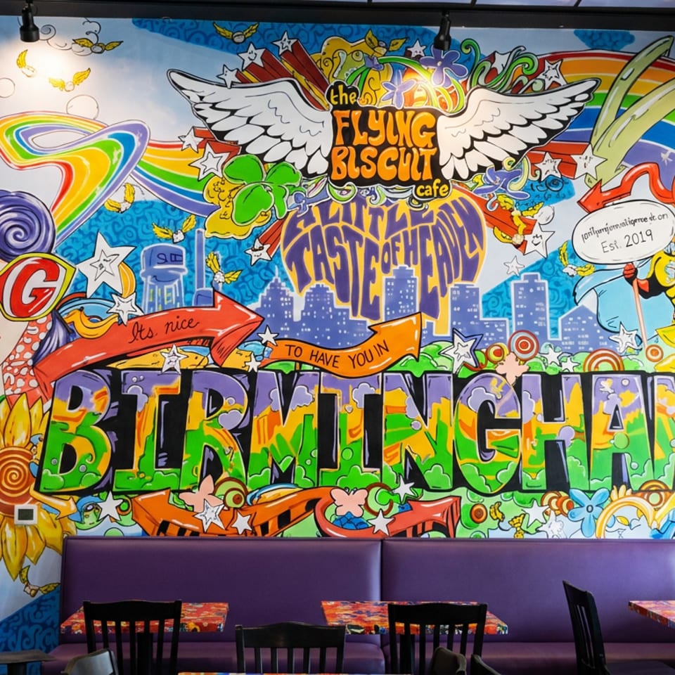 Colorful Flying Biscuit Cafe mural featuring rainbows, sunflowers, wings, city scenes, and bold lettering above purple restaurant booths.