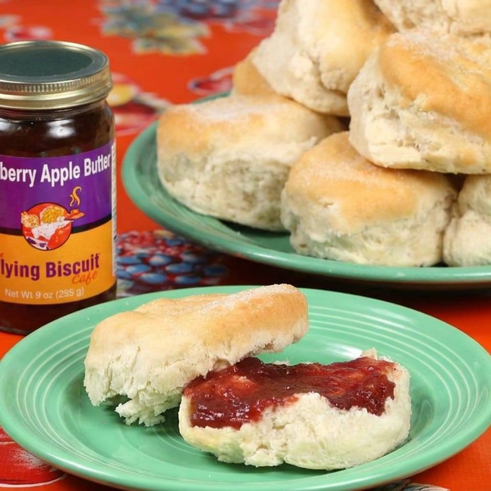 Fresh biscuits on green plates with cranberry apple butter in a jar, arranged on a colorful red fruit-patterned tablecloth.